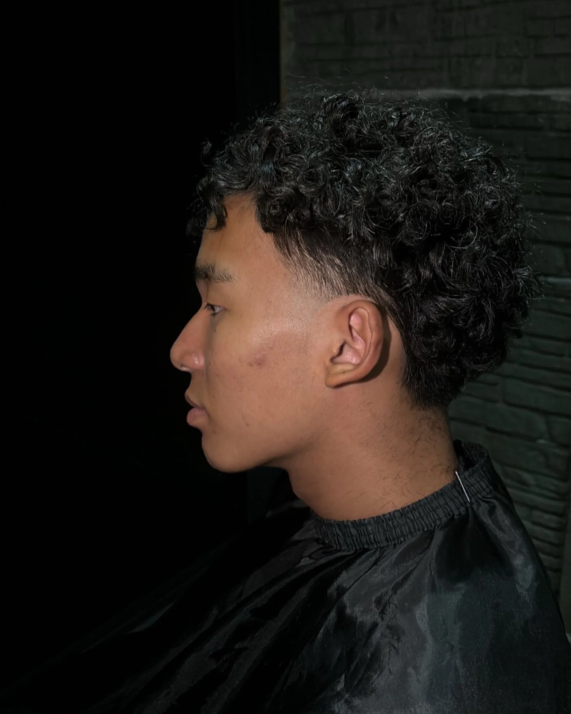 Defined curls on top, cut short around the ear, against a dark brick wall.